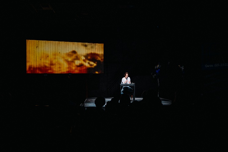dunkles Bild aus dem Zuschauerraum, auf der Bühne eine beleuchtete Person